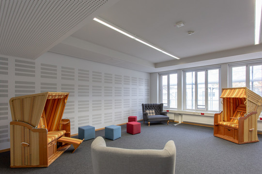 Ein Raum mit Teppichboden und Fensterfront. 4 große Sitzgelegenheiten und 4 kleine Sitzgelegenheiten.