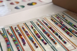 Several rows of colorful letter beads arranged on a white sheet with colored lines, next to a brown cardboard box with white paper pieces.