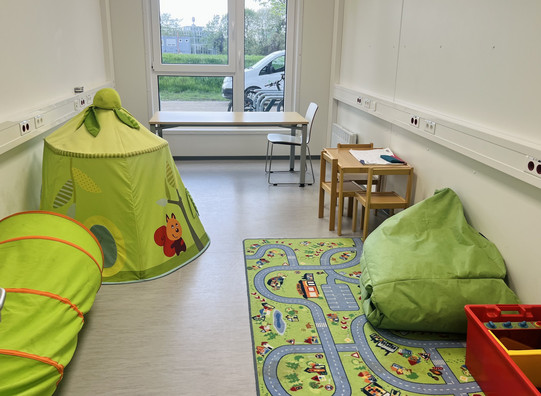 A room is shown which has a window at the end with a view of green bushes. On the floor of the room is a children's play carpet with colorful streets dot on this carpet is a beanbag and a box of children's toys in Furthermore, on the left side is a tunnel tube for small children to crawl through and a children's tent. At the back of the room is a children's table with children's chairs and another chair.