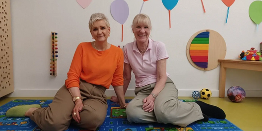 Two women sitting on a colorful play mat in a room decorated with colorful balloon shapes on the wall and toys in the background
