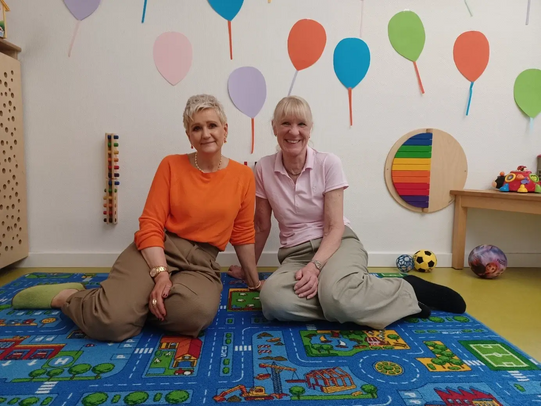 Zwei Frauen sitzen auf einem bunten Spielteppich in einem Raum mit bunten Luftballon-Dekorationen an der Wand und Spielzeug im Hintergrund