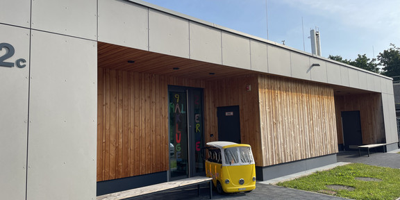 The picture shows a modern building with a combination of gray and wood-clad facades. The building is marked with the house number “2 C”. The entrance area is marked by a large glass front, which is inscribed with colorful letters forming the word “9x kluge Zwerge”. In front of the entrance is a small yellow VW bus in which small children can sit and be pushed around. The area around the building consists of a paved path and a small green lawn area, which gives the overall picture an inviting atmosphere. A clear blue sky stretches above the building.