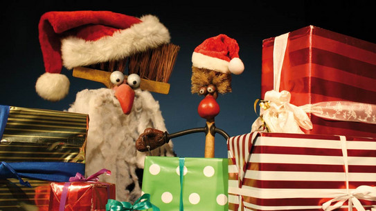 Zwei Putzutensilien mit Weihnachtsmützen, eines mit Bart und großen Augen, umgeben von bunten Geschenkpaketen.
