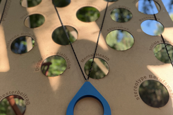 Wooden board with multiple round cutouts and a blue teardrop-shaped pendant on a string holding a pink disc with the text 'FLINTA'