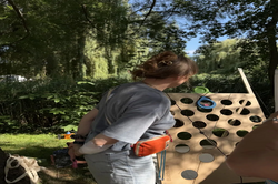 A person wearing a gray T-shirt and orange waist bag stands in front of a wooden board with many round holes in a green park.