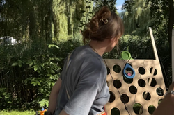 A person wearing a gray T-shirt and orange waist bag stands in front of a wooden board with many round holes in a green park.