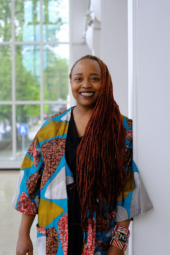 Photo of Dr Rahab Njeri, with long dreadlocks and a colourful jacket, leaning against a white wall in front of a large window