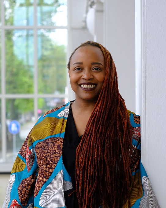 Photo of Dr Rahab Njeri, with long dreadlocks and a colourful jacket, leaning against a white wall in front of a large window