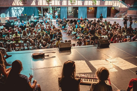View from the stage toward an audience of children and adults in a hall with an instrument and sheet music on the stage