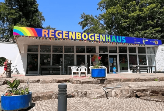 Entrance of a building with a large colorful sign reading 'REGENBOGENHAUS' and several white chairs in front