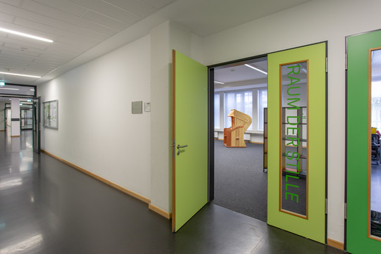 Foto des Eingangsbereichs. Die grüne Tür steht offen und man schaut in den Raum hinein und sieht einen orangenen Strandkorb. Die andere Hälfte der Tür hat ein Fenster, auf dem "Raum der Stille" steht. 