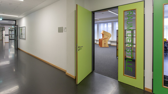 Foto des Eingangsbereichs. Die grüne Tür steht offen und man schaut in den Raum hinein und sieht einen orangenen Strandkorb. Die andere Hälfte der Tür hat ein Fenster, auf dem "Raum der Stille" steht.