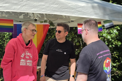 Three people under a white canopy with rainbow and transgender flags standing at a table covered with a rainbow flag and flyers.