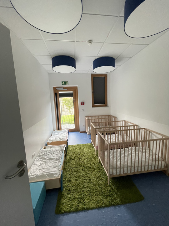 In diesem Bild ist ein heller und freundlicher Raum zu erkennen, der Schlafraum für Kleinkindern gestaltet ist. Der Raum verfügt über mehrere Gitterbetten. Ein einladender, grüner Teppich sorgt für eine gemütliche Atmosphäre. An der Wand hängen große, runde Lampen in Blau, die sanftes Licht spenden. Ein Fenster lässt Tageslicht herein und bietet einen Blick nach draußen, was den Raum noch freundlicher wirken lässt. Insgesamt vermittelt der Raum ein Gefühl von Sicherheit und Geborgenheit für Kleinkinder.