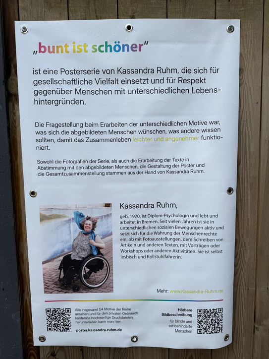 A poster with a white background and black text, featuring the colorful headline 'bunt ist schöner' at the top center. Below is an explanatory text about a postcard series by Kassandra Ruhm, which advocates for social diversity and respect for people with different life backgrounds. In the lower section, there is a photo of a person in a wheelchair sitting outdoors on a path, waving with one hand. To the right of the photo is a brief biography of Kassandra Ruhm. At the bottom edge, two QR codes and a web address are visible.