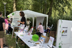 Outdoor booth with informational materials on a table under trees, people standing and sitting nearby, banner with the word 'AWARENESS'.
