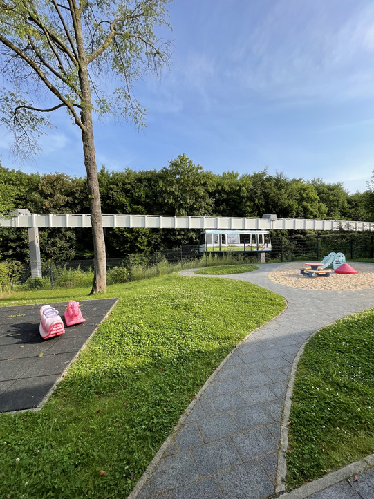 Auf dem Bild sieht man das Außengelände der Großtagepflegestellen am Campus Süd, welches eine grüne Wiese und einen Sandkasten umfasst. Im Garten stehen mehrere Spielgeräte, wie kleine Wippsitze und eine Rutschen. Im Hintergrund erkennt man eine vorbeifahrende H-Bahn. Der Himmel ist klar und blau, was auf einen sonnigen Tag hindeutet.