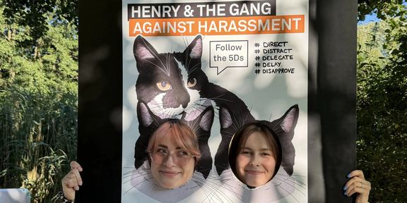 Two people hold a large poster in front of themselves, covering their bodies and showing only their faces through two circular cutouts. The poster features two cat heads with black fur and white markings at the top. Large text at the top reads: 'HENRY & THE GANG AGAINST HARASSMENT'. Below, a box contains the text: 'Follow the 5Ds'. To the right, five terms are listed: 'DIRECT', 'DISTRACT', 'DELEGATE', 'DELAY', 'DOCUMENT'. At the bottom, the poster displays: 'SchuDs' and underneath: 'Zentrale Beratungsstelle zum Schutz vor Diskriminierung und sexualisierter Gewalt'. Trees and sunlight are visible in the background.