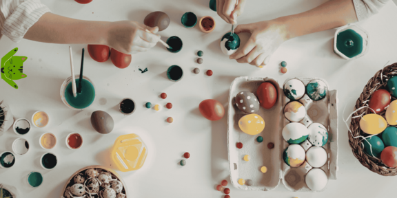 Two hands painting colorful Easter eggs on a table with paint pots and painted eggs in an egg carton and a basket