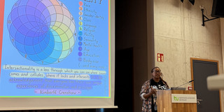 Dr. Rahab Njeri steht an einem Rednerpult in einem Hörsaal vor einer Leinwand mit einer farbigen Grafik zum Thema Intersektionalität