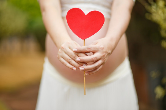 A woman with a baby bump is shown. She holds a paper heart in front of her belly.