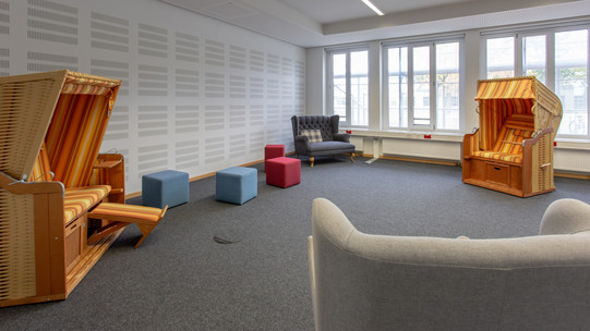 Ein Foto mitten im Raum der Stille. Man sieht zwei orangene Strandkörbe, ein Zweisitzer-Sofa, mehrere blaue und rote Sitzhocker und die Lehne eines Sessels, von hinter dem das Foto aufgenommen wurde.