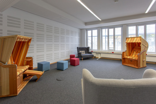 Ein Foto mitten im Raum der Stille. Man sieht zwei orangene Strandkörbe, ein Zweisitzer-Sofa, mehrere blaue und rote Sitzhocker und die Lehne eines Sessels, von hinter dem das Foto aufgenommen wurde.