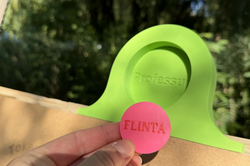 Hand holding a round pink sticker with the text 'FLINTA' in front of a green clip labeled 'Professur' attached to brown paper outdoors with a blurred forest background.
