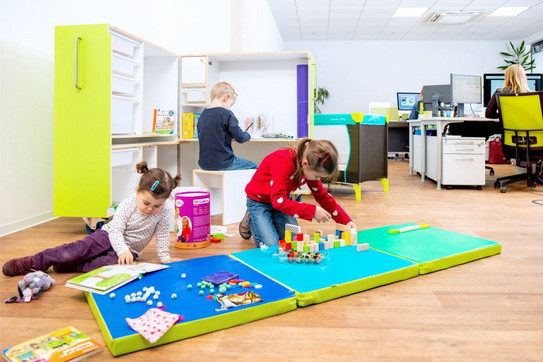 Zwei Mädchen spielen mit Spielsachen auf einer Gymnastikmatte. Ein Junge mal auf dem Schreibtisch der KidsBox