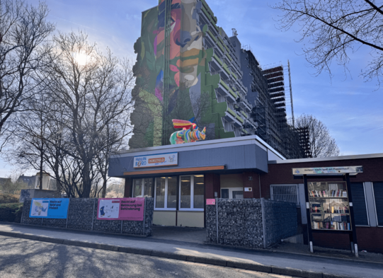 Multi-story residential building with large colorful mural and small building with signs and a bookshelf in front