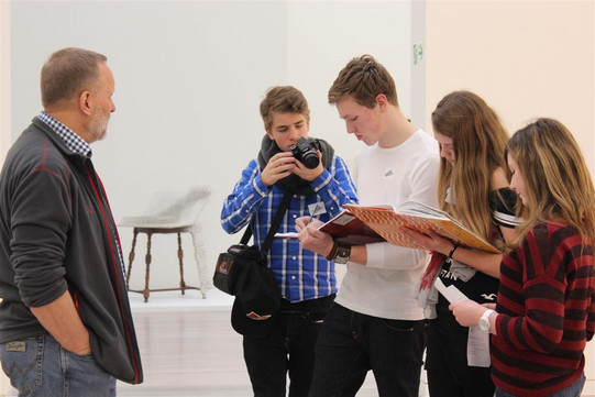 A man stands on the left, hands in his pockets, on the left are three people bent over a flyer, another is photographing the group 