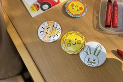 Four round, hand-painted children's motifs featuring a sun, bee, and rabbit lie on a wooden table next to two red wax crayons and an orange cup.