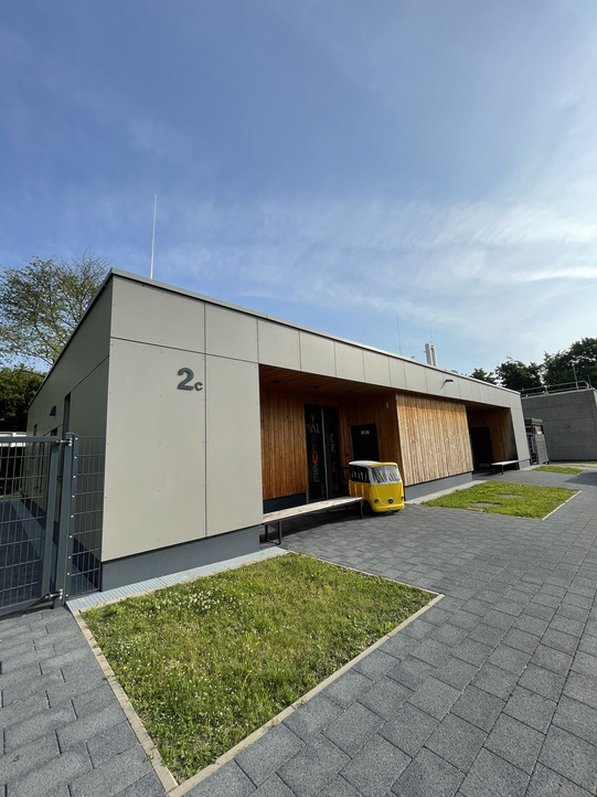 Das Bild zeigt ein modernes Gebäude mit einer klaren, minimalistischen Architektur. Die Fassade besteht aus glatten, hellgrauen Paneelen und einem großen Bereich aus Holz, der eine warme Note hinzufügt. An der Vorderseite des Gebäudes ist die Nummer "2c" deutlich sichtbar angebracht. Vor dem Eingang steht ein kleiner gelber Wagen, der einen fröhlichen Kontrast zur neutralen Farbpalette des Gebäudes bildet. Im Hintergrund sind einige Bäume und weitere Strukturen zu erkennen, während der Himmel leicht bewölkt ist und das Licht sanft auf die Szene fällt. Der gepflegte Außenbereich umfasst sowohl Pflastersteine als auch grüne Rasenflächen.