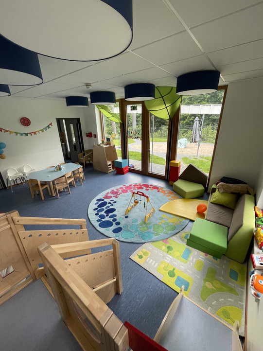 In diesem Raum sieht man eine freundliche und einladende Umgebung, die für Kinder gestaltet wurde. Der Raum ist hell und gut beleuchtet, mit großen Fenstern, die einen Blick auf den Außenbereich bieten. Auf dem Boden liegt ein bunter Teppich mit verschiedenen Formen und Farben, der zum Spielen einlädt.   Es gibt gemütliche Sitzgelegenheiten in Form von grünen Sofas und Kissen sowie einige Spielzeuge, die auf einem Regal oder Tisch verteilt sind. In einer Ecke steht ein kleiner Tisch mit Stühlen, der für kreative Aktivitäten genutzt werden kann. Die Wände sind mit bunten Dekorationen geschmückt, die eine fröhliche Atmosphäre schaffen.  Die Decke ist mit farbenfrohen Lampen ausgestattet, die dem Raum zusätzliches Licht verleihen. Insgesamt vermittelt der Raum eine freundliche und spielerische Stimmung, ideal für Kinder zum Lernen und Entdecken.