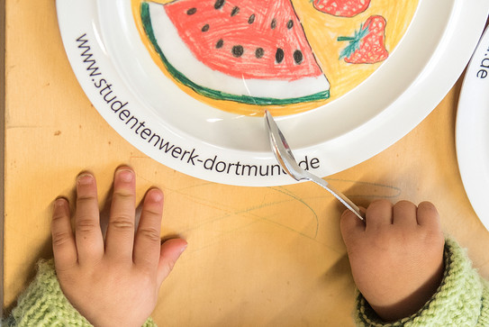 Child's hands are holding a fork next to a plate.