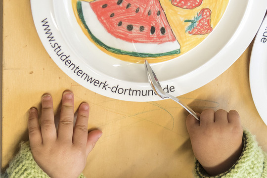 Child's hands are holding a fork next to a plate.