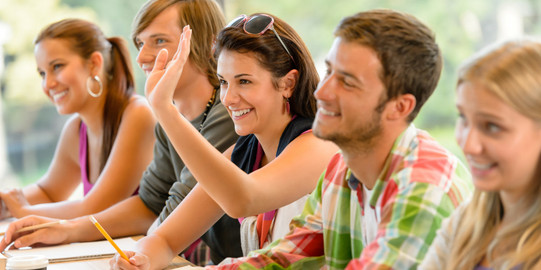 Schülergruppe Zwei Jungen und zwei Mädchen sitzen neben einander an einem Tisch im Klassenraum und heben die Hand.